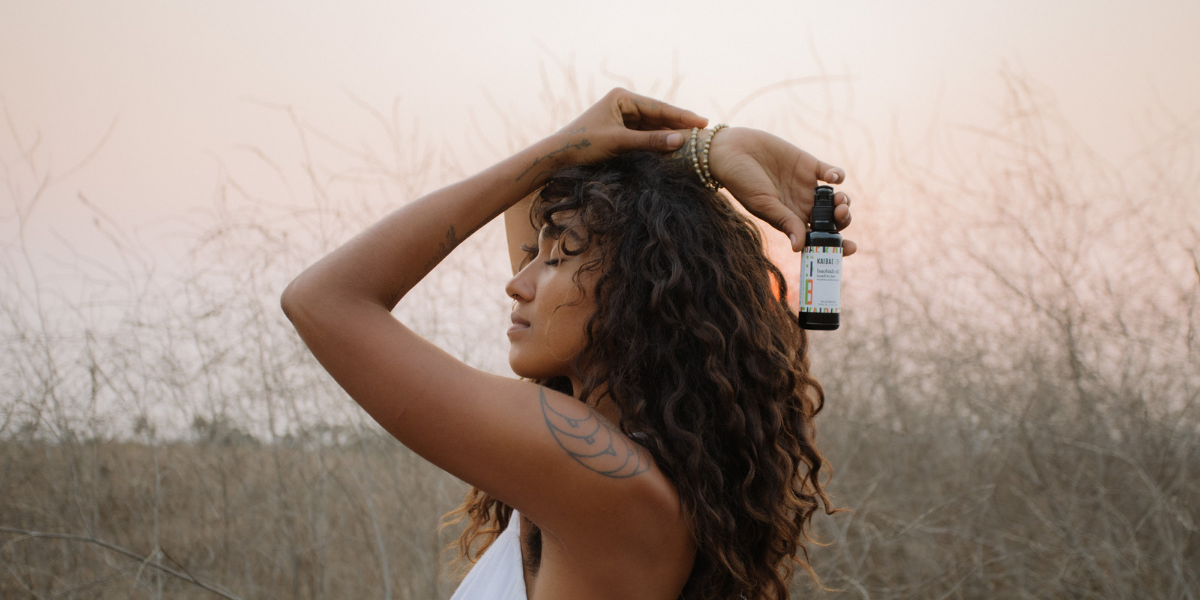 Young Woman standing in a field of wild grasses with a bottle of organic wild harvested Baobab Oil nourishing for hair and skin from head to toe I KAIBAE