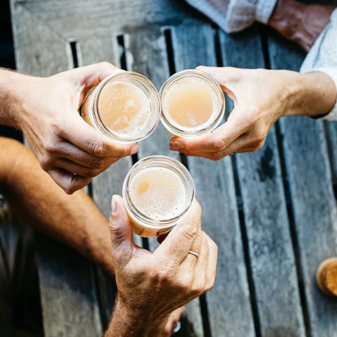Three hands hold glasses filled with a light-colored drink, infused with KAIBAE's baobab powder: prebiotic super fiber, and are raised in a toast over a wooden table. The scene conveys camaraderie and celebration while subtly highlighting the benefits of gut health and prebiotic fiber.