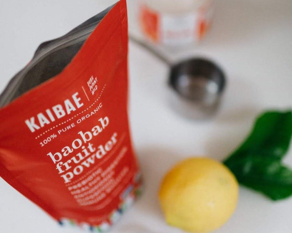 A red package of KAIBAE baobab powder: prebiotic super fiber, known for its gut health benefits, rests on a white surface. Positioned nearby are a lemon, a metal measuring cup, and two green leaves, emphasizing its natural origins.