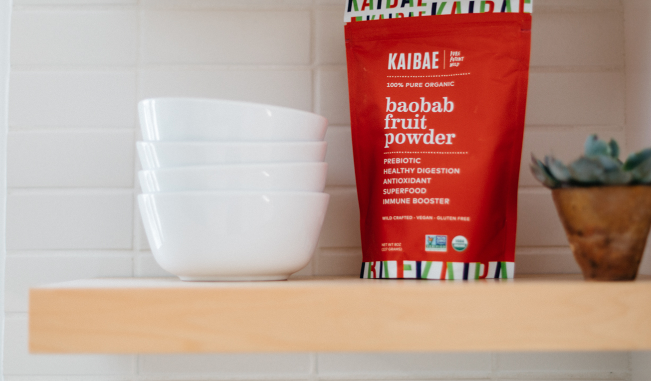 A KAIBAE baobab powder bag, brimming with prebiotic fiber for gut health, is showcased on a wooden shelf beside four neatly stacked white bowls. A small succulent plant adorns the right side of the shelf, set against a backdrop of white tiled walls.