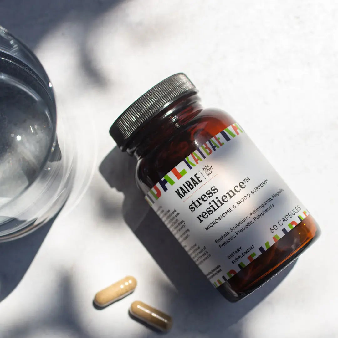 stress resilience by KAIBAE , a dietary supplement lying on a white counter with a 2 capsules next to the amber glass bottle, lying in the light of the sun next to a glass of water