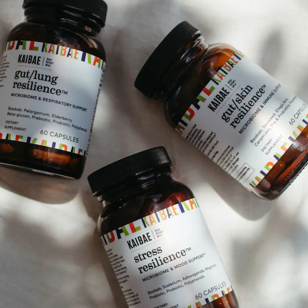 Three bottles of kaibae supplements labeled for gut/lung resilience, stress resilience, and microbiome support, scattered on a surface with sunlight casting shadows.