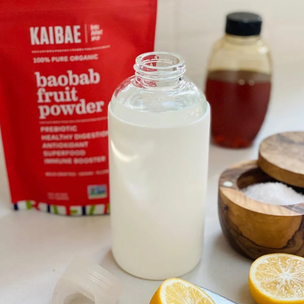 A frosted glass bottle with a creamy beverage next to a red pack of KAIBAE baobab powder, rich in prebiotic fiber for gut health, a bowl of salt, and sliced lemons. A bottle of honey is in the background.