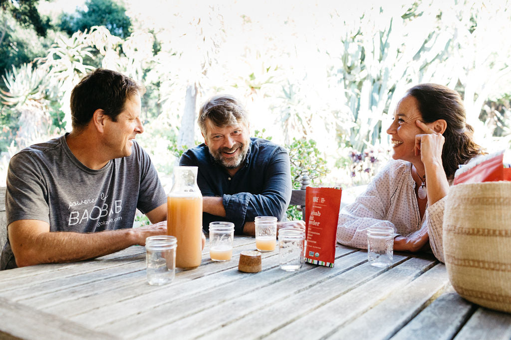 KAIBAE cofounders Thomas Cole, Dr. Luc Maes and Barbara Berger Maes sitting at a table, enjoying each other and drinking a pitcher of prebiotic Baobab lemonade. 