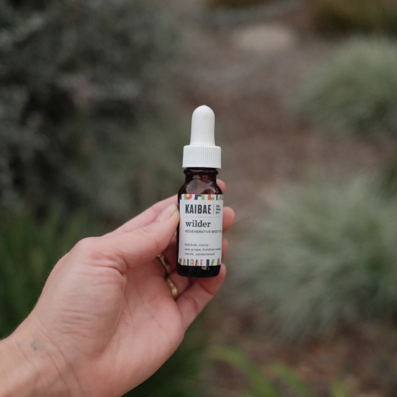 A hand holds a small bottle with a dropper lid labeled "KAIBAE wilder mini," set against a blurred natural background of greenery, containing luxurious body oil infused with wild plant oils such as Sandalwood and Neroli.