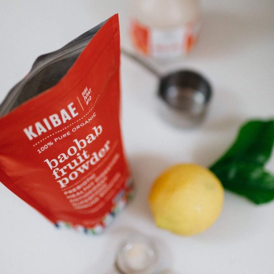 A red package of KAIBAE baobab powder: prebiotic super fiber, known for its gut health benefits, rests on a white surface. Positioned nearby are a lemon, a metal measuring cup, and two green leaves, emphasizing its natural origins.