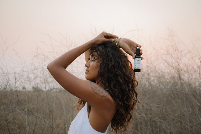 Woman standing in a field of wild grass holding a bottle of KAIBAE Baobab oil for use from hair to toes. 