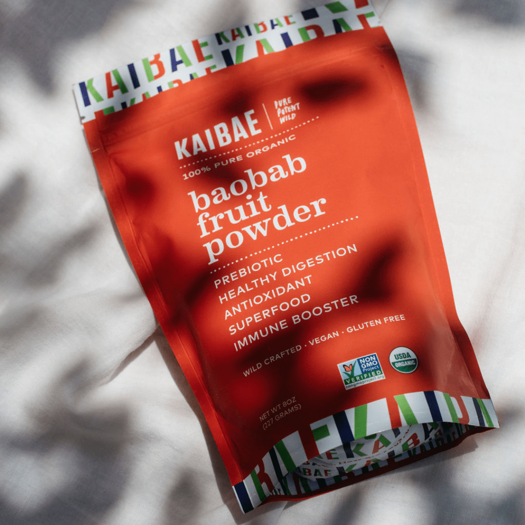 A red pouch labeled "KAIBAE Baobab Powder" lies on a white surface with shadows of leaves cast over it. The packaging promotes it as a prebiotic, healthy for digestion and gut health, antioxidant-rich, superfood, immune booster, vegan, and gluten-free.