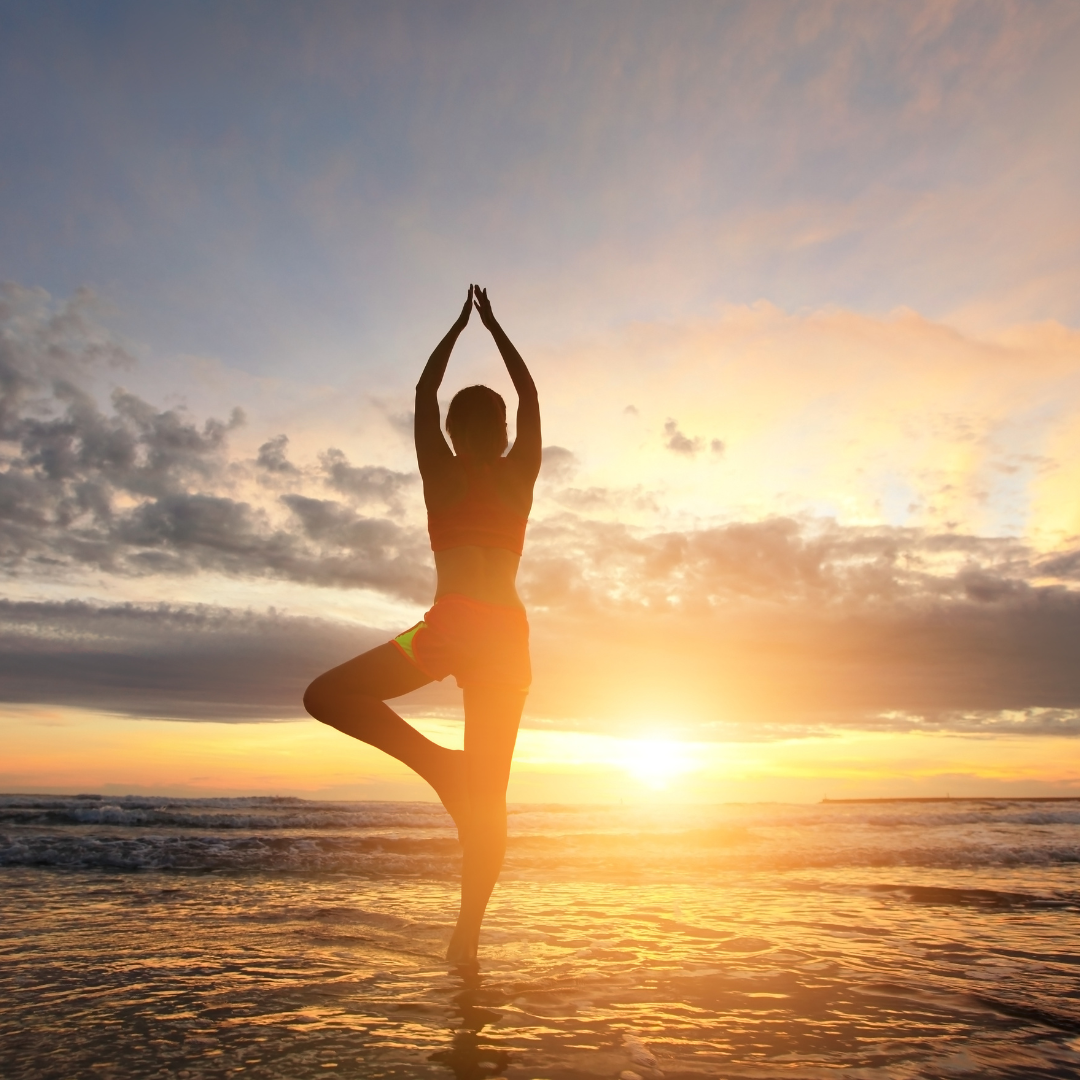 A person stands on one leg in a yoga pose with arms raised above their head on a beach during sunset. The sky is a blend of orange, yellow, and blue hues as the sun is low on the horizon, casting a beautiful reflection on the water—perfect for soothing the stress response with KAIBAE's Resilience Series.