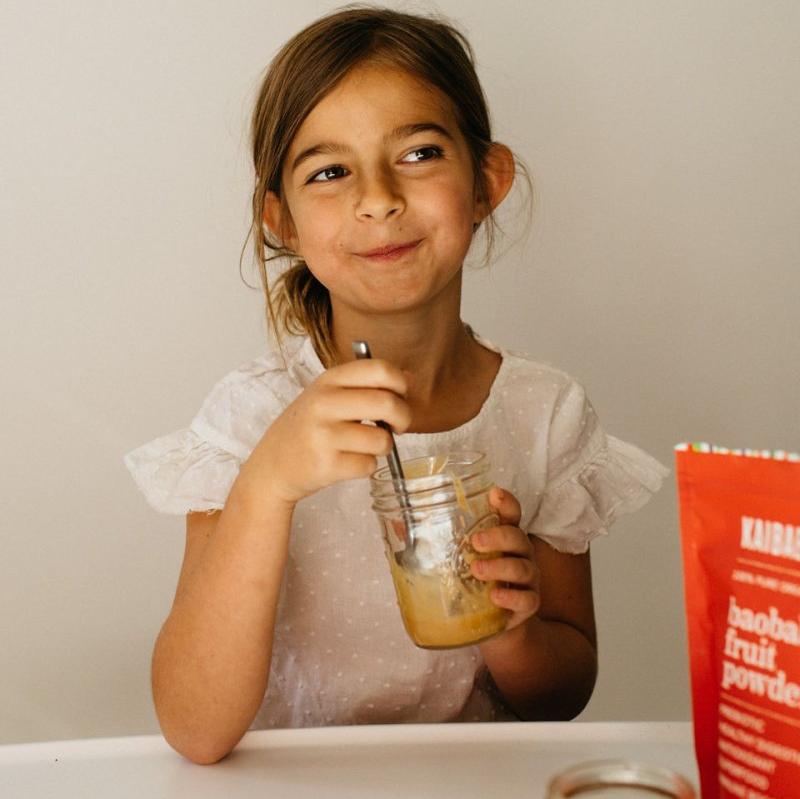 A young girl in a white dress is sitting at a table, smiling while holding a jar with a spoon. Beside her on the table is a red packet of KAIBAE's baobab powder: prebiotic super fiber, known for supporting gut health. The setting is minimalistic with a light background.