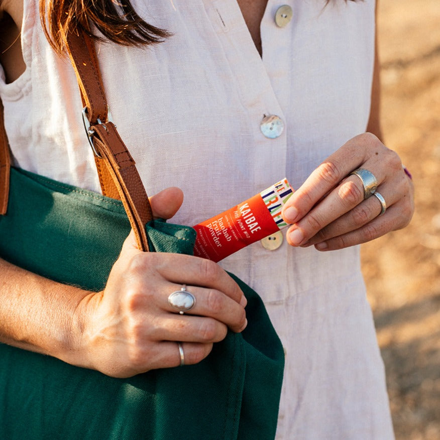 A person wearing a light sleeveless top places a box of baobab powder, 5 single sticks from KAIBAE, into a green bag with brown leather straps. The person is wearing rings on both hands, including a large white stone ring. The baobab powder box has a colorful label and is red.