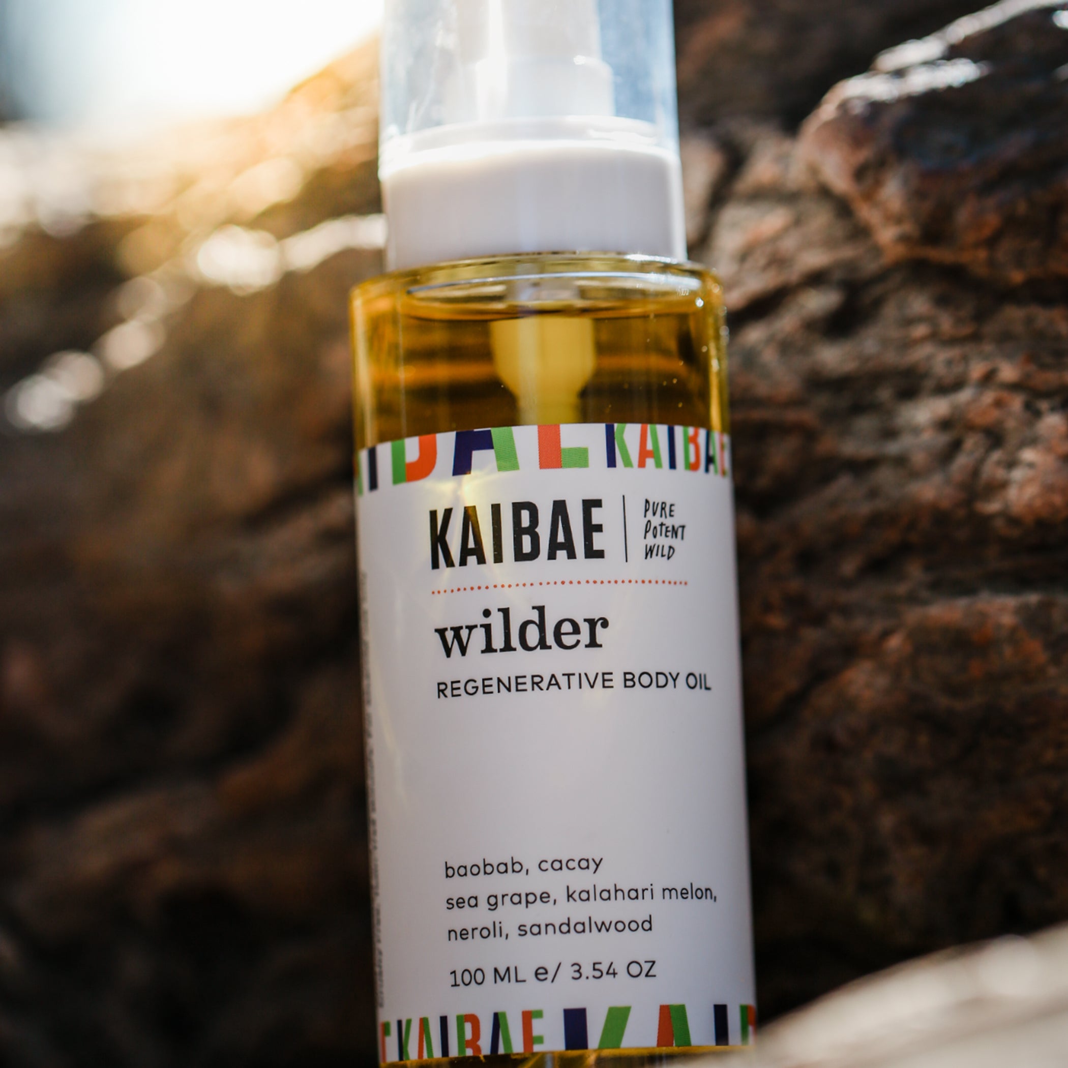 A bottle of kaibae "wilder" regenerative body oil, highlighted by the sunset, showing ingredients like baobab and cacay, seagrape, kalahari melon seed oil, neroli and sandalwood on its label, against a rocky background.